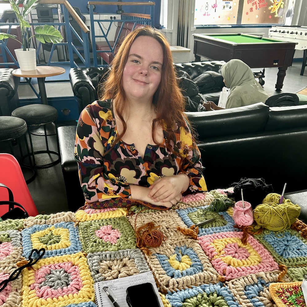 Person with long ginger hair sitting at a table with handmade crochet squares of different colours