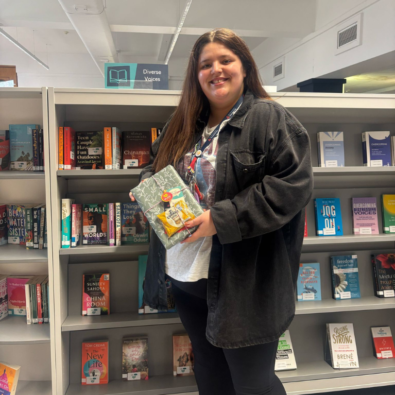 Person with shoulder length dark brown hair stood in a library holding two books smiling at the camera