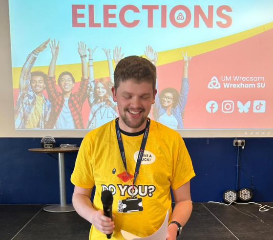 Person with short brown hair wearing a yellow tshirt holding a microphone
