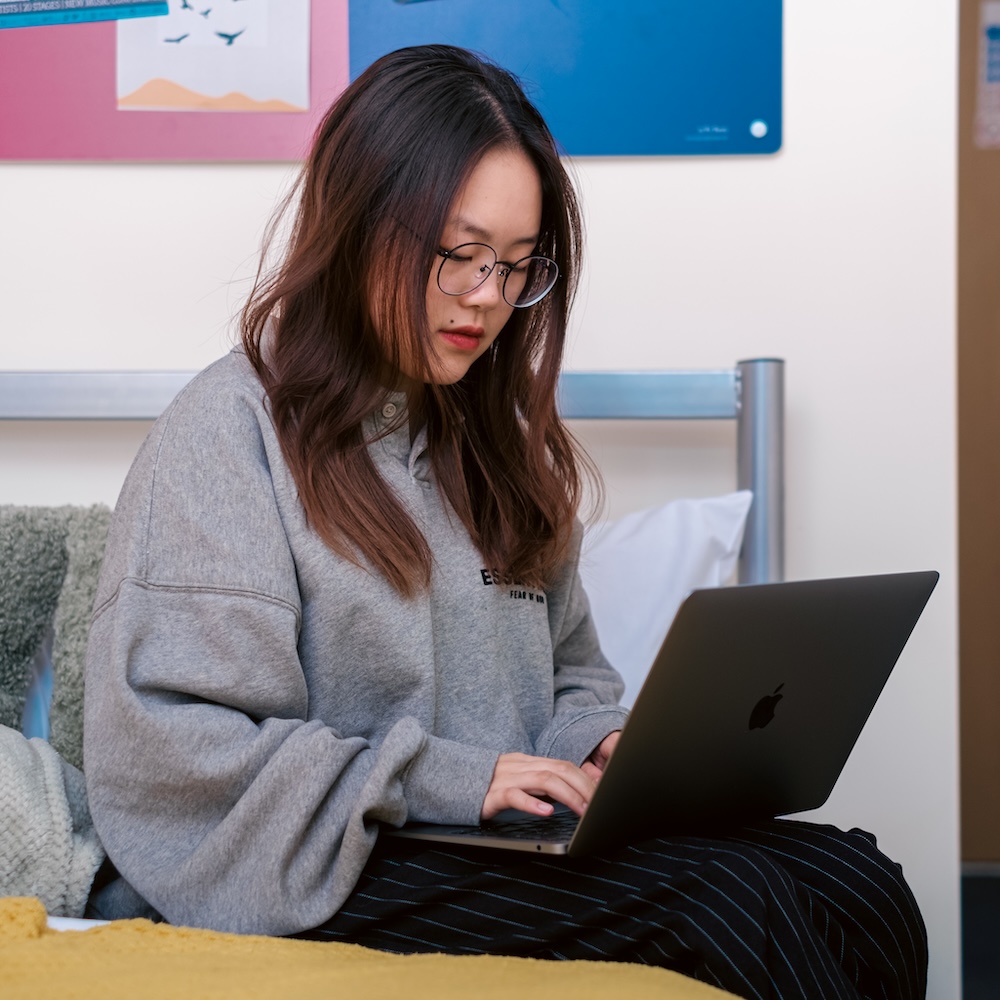 Person with long black hair sitting on their bed with a laptop typing